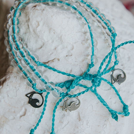 A limited edition set of 4Ocean 4OCEAN BRACELET WORLD OCEAN DAY LIMITED EDITION, with small silver fish hook charms and clear round beads, made from recycled materials, placed on a white stone surface.