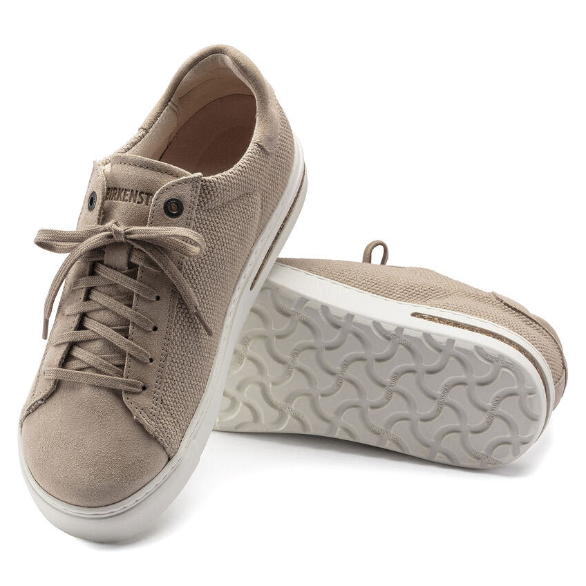 A pair of beige fabric sneakers with white rubber soles; one shoe is upright while the other is resting on its side, showcasing the sole&#39;s tread pattern. Inspired by the classic BIRKENSTOCK BEND SANDCASTLE CANVAS - WOMENS, these women&#39;s shoes from Birkenstock feature an anatomically shaped cork-latex footbed for ultimate comfort.