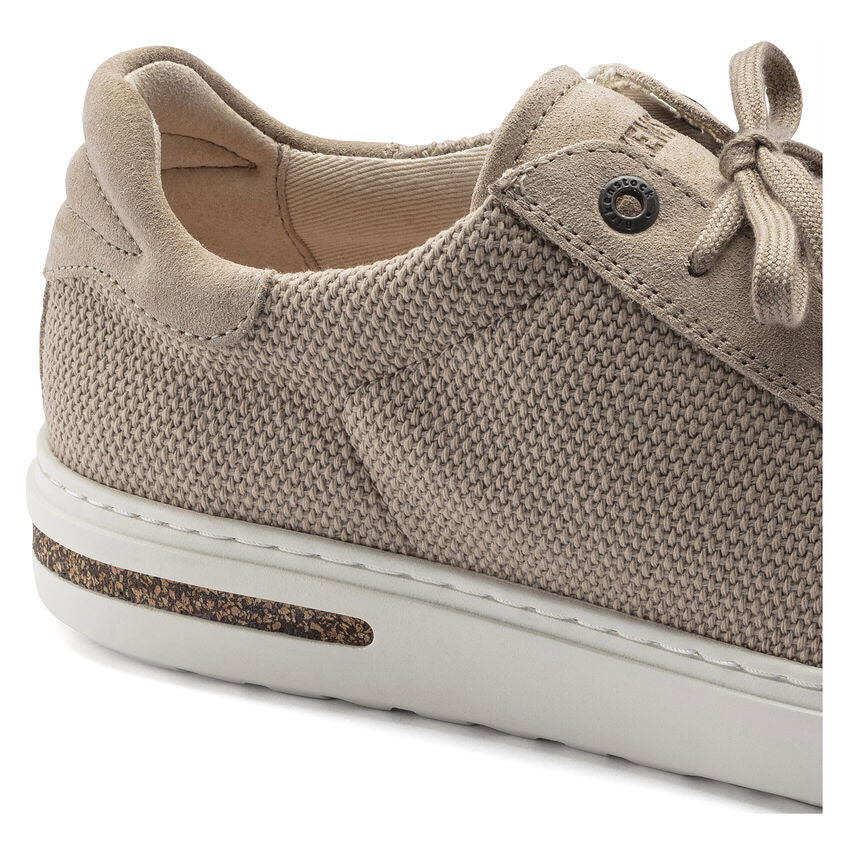 Close-up of a beige Birkenstock BIRKENSTOCK BEND SANDCASTLE CANVAS - WOMENS sneaker with a knitted upper, white sole, and brown detailing near the heel. The shoe features laces, a padded heel collar, and an anatomically shaped footbed for optimal shock absorption.