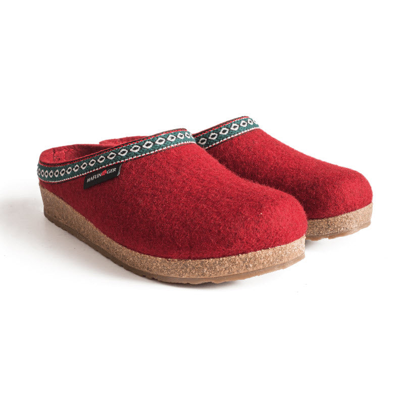 A pair of red woolen clogs with a decorative trim around the opening, featuring cork and latex midsoles and a label on the side of each shoe, such as the HAFLINGER GZ CHILI - WOMENS by Haflingers.