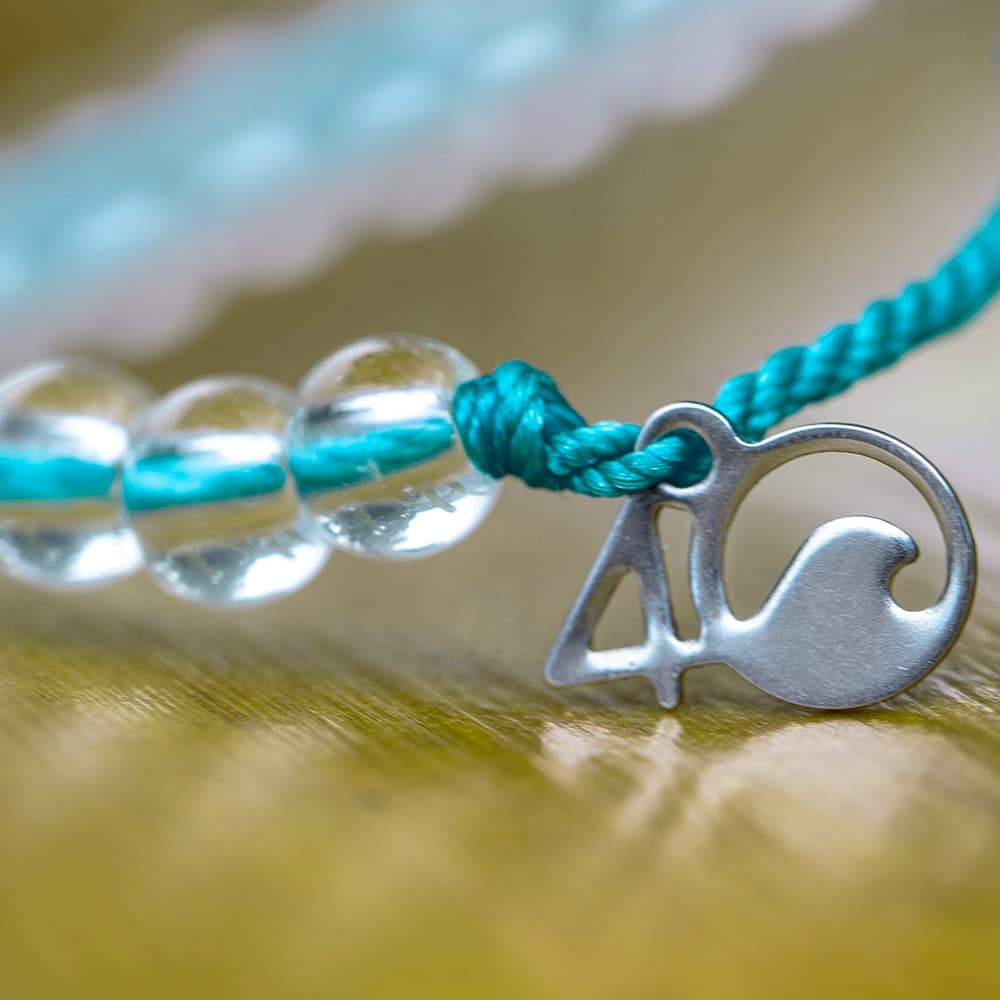 Close-up of a 4OCEAN BRACELET WHITE SIDED DOLPHIN by 4Ocean, with transparent beads threaded onto a turquoise string, featuring a small metal charm shaped like the number 4 and a wave, raising awareness about plastic pollution.