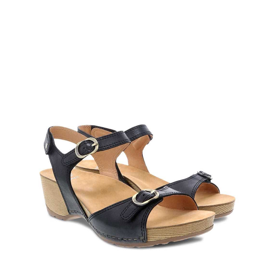 The DANSKO TRICIA BLACK MILLED BURNISHED - WOMENS by Dansko are a pair of black wedge sandals featuring leather uppers, ankle straps with metal buckles, and tan wooden soles that offer all-day comfort.