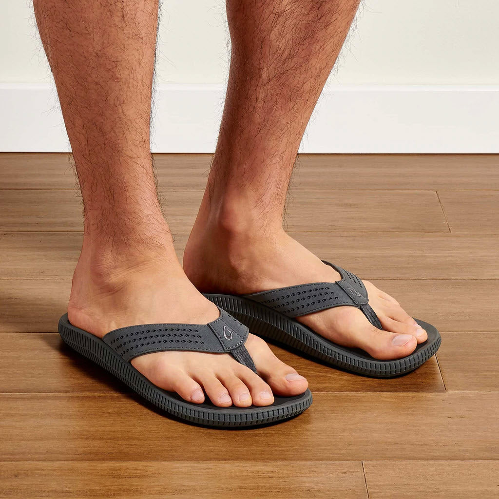 A person wearing Olukai Ulele Dark Shadow sandals stands on a hardwood floor near a white wall. Only the lower legs and feet are visible.