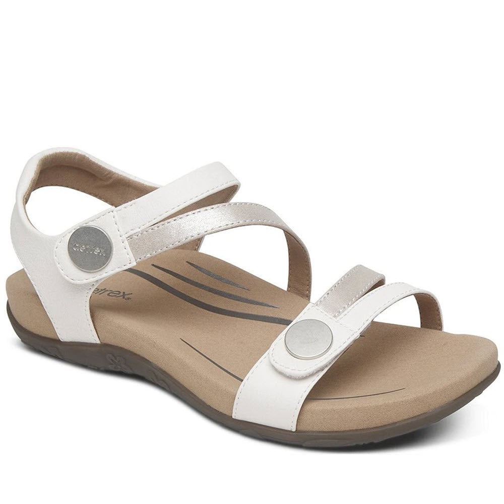 Aetrex AETREX JESS WHITE - WOMENS is a white, adjustable sandal with a beige inner sole and two adjustable Velcro straps on top, featuring silver circular accents and enhanced arch support.