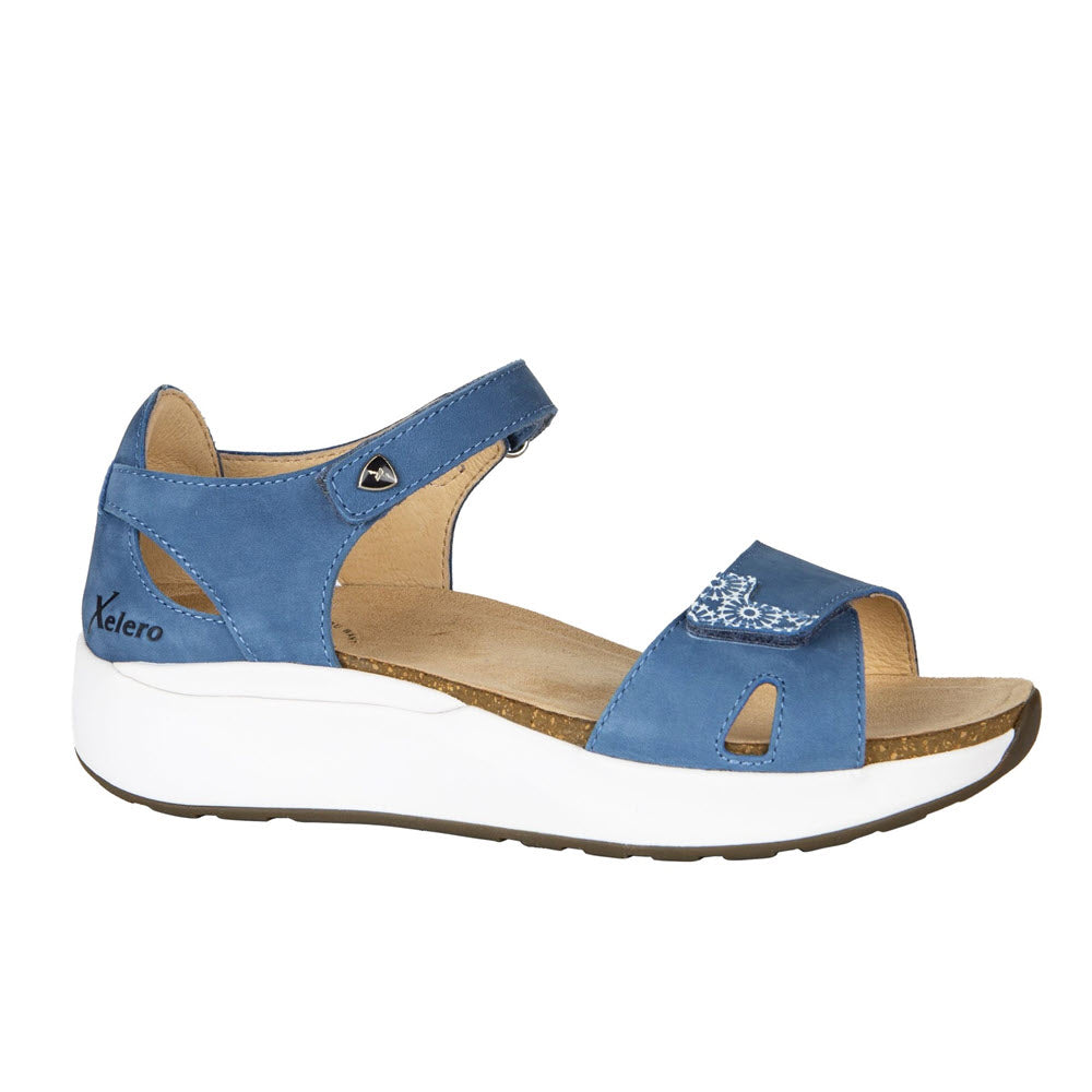 The Xelero Santorini Denim Nubuck for women is a blue orthopedic sandal featuring an open toe, adjustable straps, and decorative white stitching on the front strap. Its thick, slip-resistant white sole includes a logo near the heel and provides excellent stability and cushioning for all-day comfort.