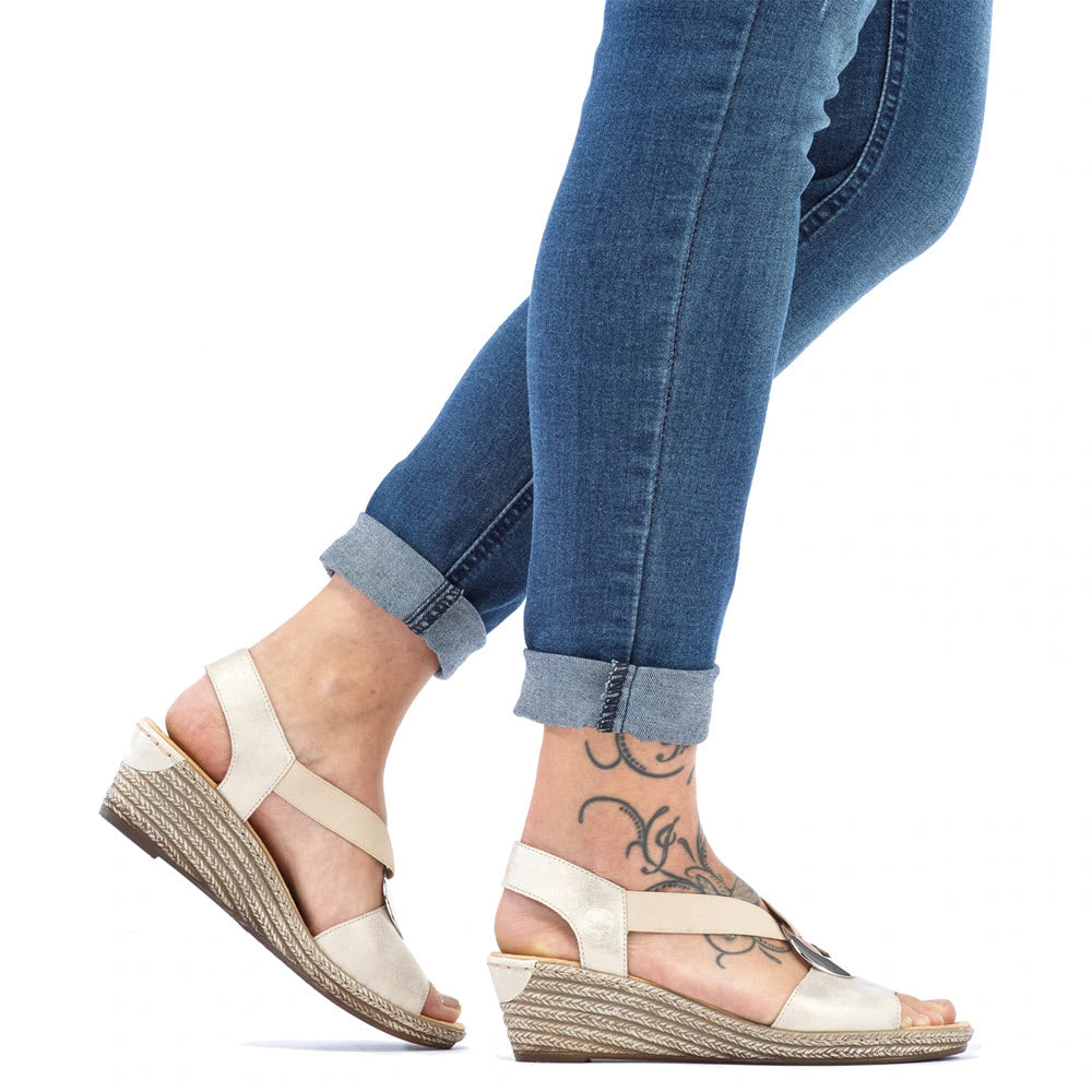 Person wearing rolled-up jeans and Rieker RIEKER SLINGBACK WEDGE WITH ORNAMENT CHAMPAGNE - WOMENS, displaying a foot tattoo, shown walking.