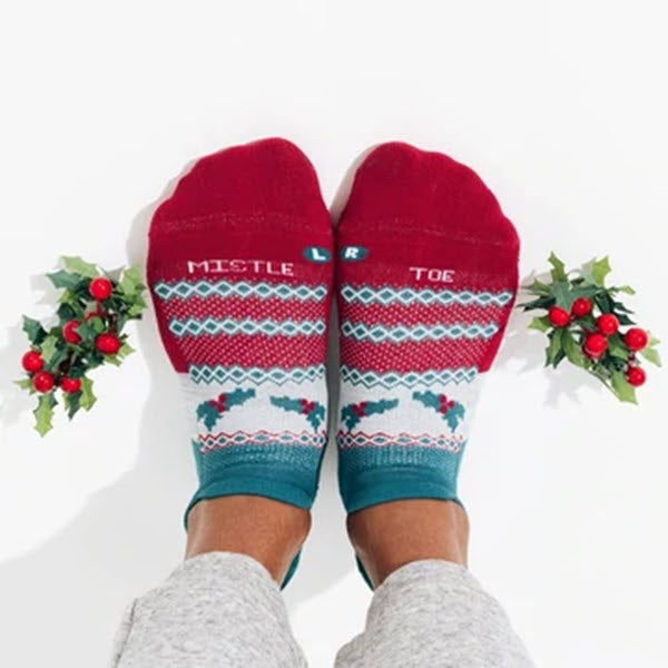 Feet wearing red and blue holiday-themed socks with &quot;MISTLE TOE&quot; written on them, surrounded by small sprigs of holly. These festive beauties, designed with a light cushion for added comfort, could easily be mistaken for Feetures No Show Socks Miseltoe - Womens style but perfect for showing off during the holidays.