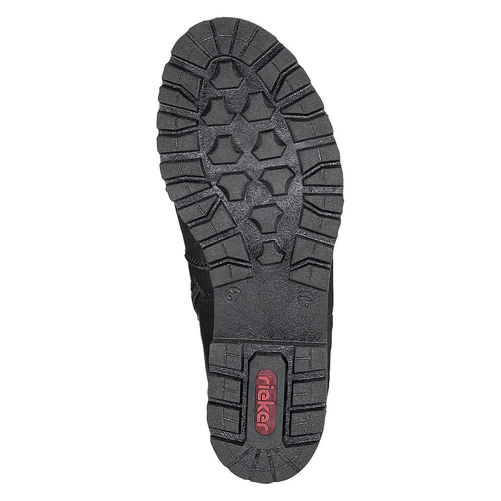 The image shows the sole of a black RIEKER LUG SOLE CHELSEA WITH ZIP BLACK - WOMENS with rugged treads, featuring the brand name &quot;Rieker&quot; in red near the heel. The design promises enhanced wintertime walkability.