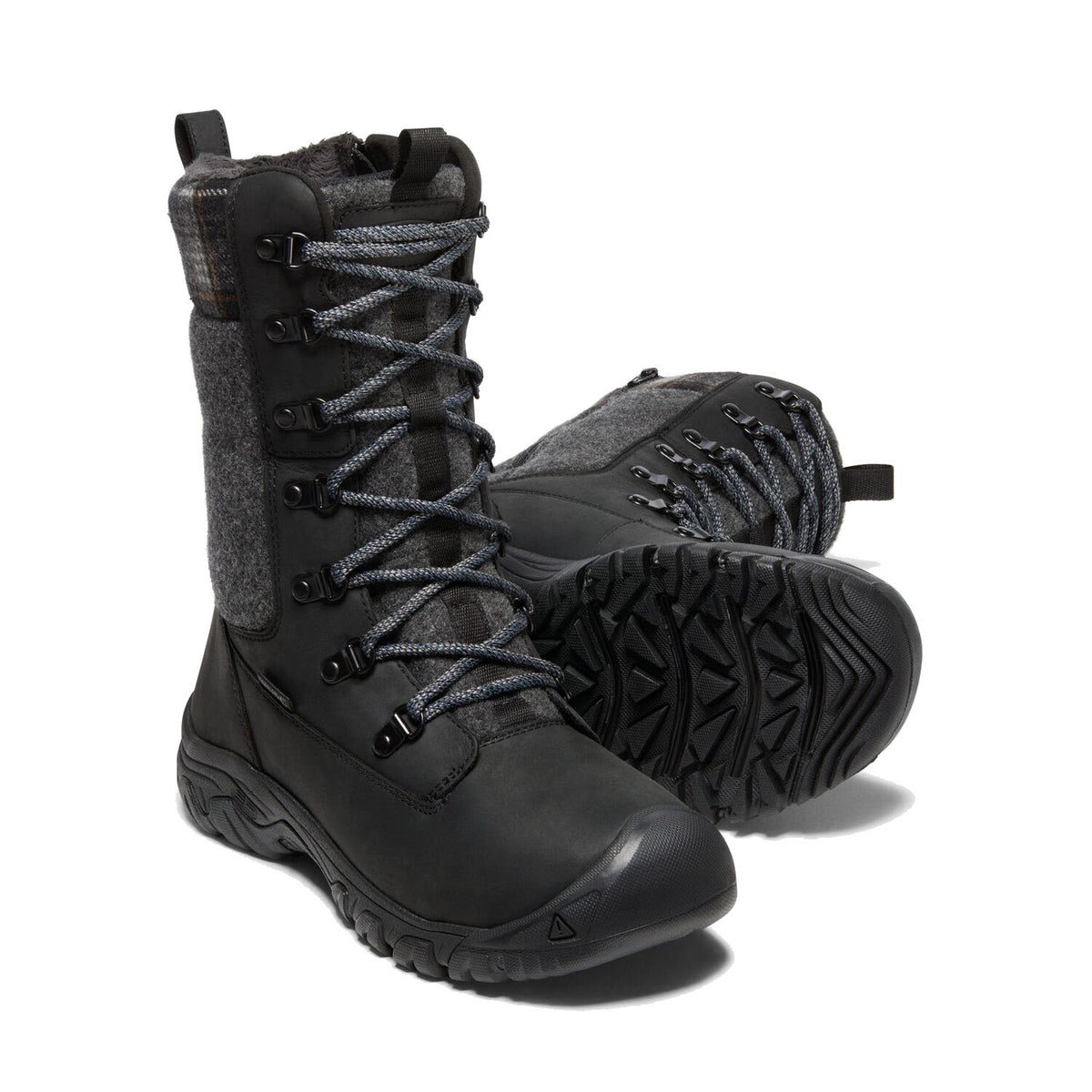 A pair of KEEN GRETA TALL BOOT WP BLACK - WOMENS from Keen, featuring thick soles and lace-up fronts for optimum cold-weather traction, with one boot standing upright and the other lying on its side.