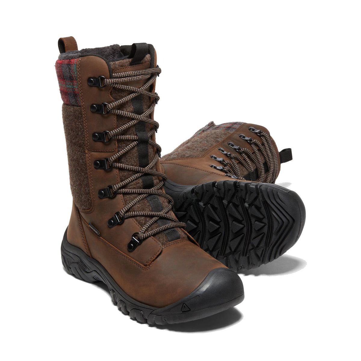 Introducing the KEEN GRETA TALL BOOT BROWN - WOMENS: A stylish pair of brown leather waterproof hiking boots from Keen, featuring black lug soles for excellent traction, plaid and fabric detailing for a unique look, and a lace-up front for a secure fit. One boot stands upright, while the other lies on its side to showcase the intricate tread pattern.