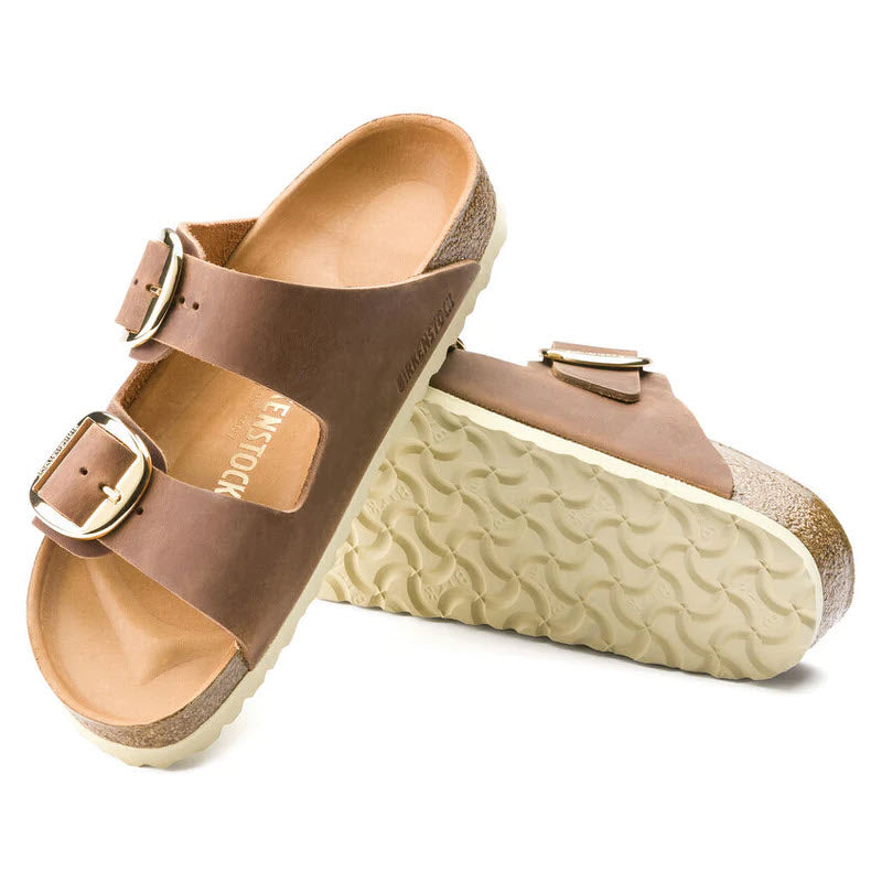 A pair of Birkenstock BIRKENSTOCK ARIZONA BIG BUCKLE COGNAC OILED - WOMENS sandals, featuring a two-strap design with metal buckles and contoured cork footbeds, positioned with one sandal leaning on the other.