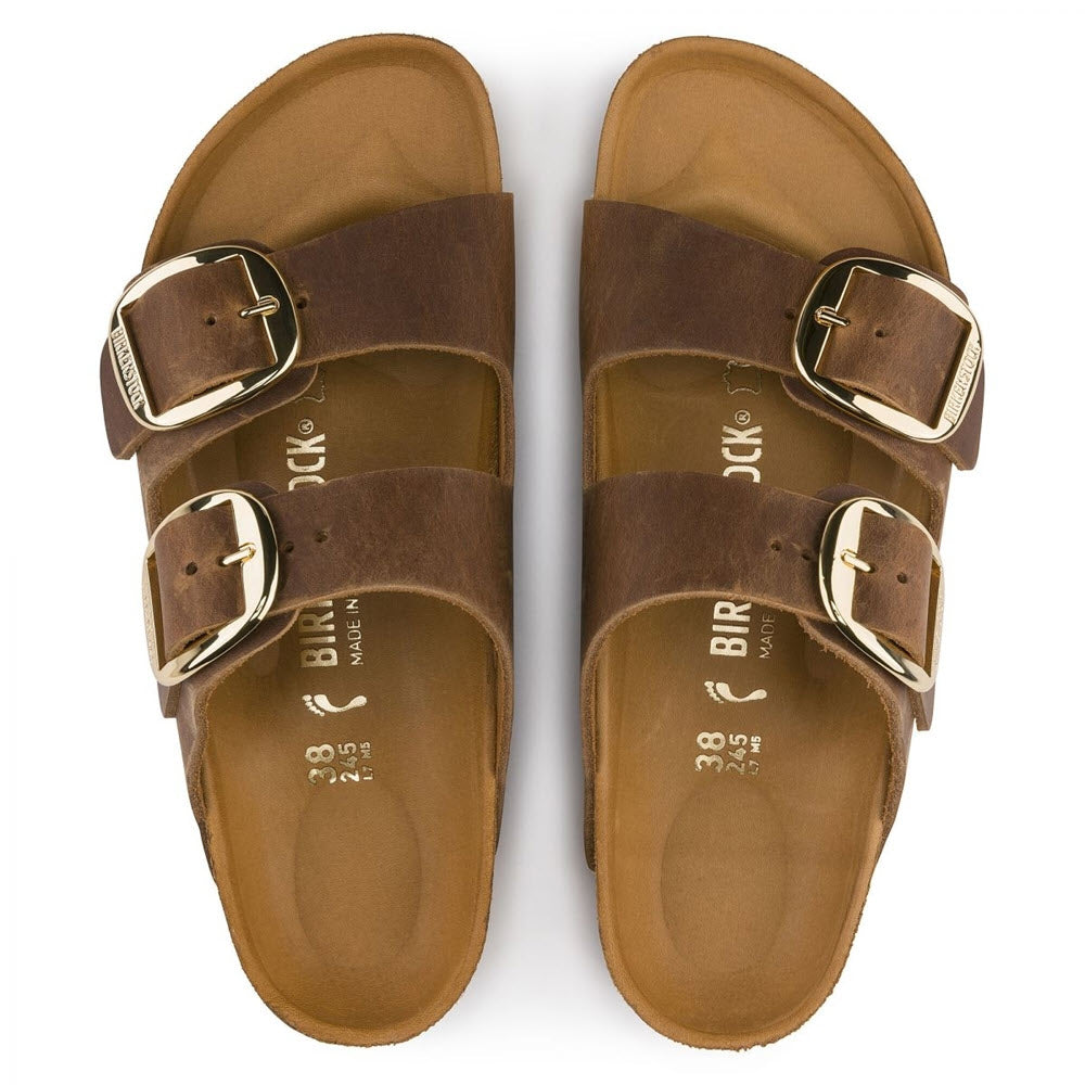 A pair of brown Birkenstock BIRKENSTOCK ARIZONA BIG BUCKLE COGNAC OILED - WOMENS sandals with a two-strap design and gold buckles on each.