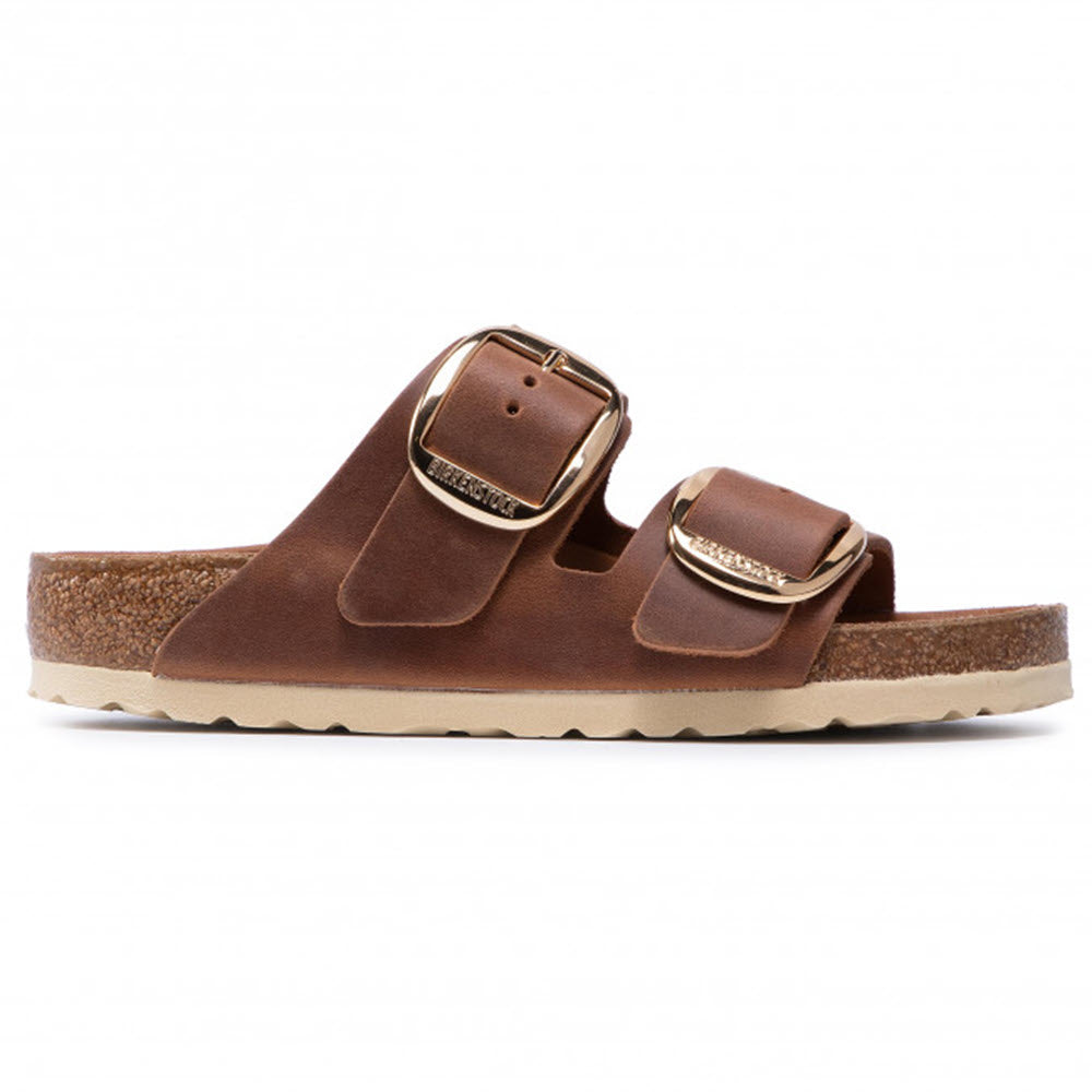 Brown leather sandal with a two-strap design featuring gold buckles and a cork footbed. The sole is light-colored and textured for grip. This BIRKENSTOCK ARIZONA BIG BUCKLE COGNAC OILED - WOMENS model from Birkenstock combines both style and comfort effortlessly.