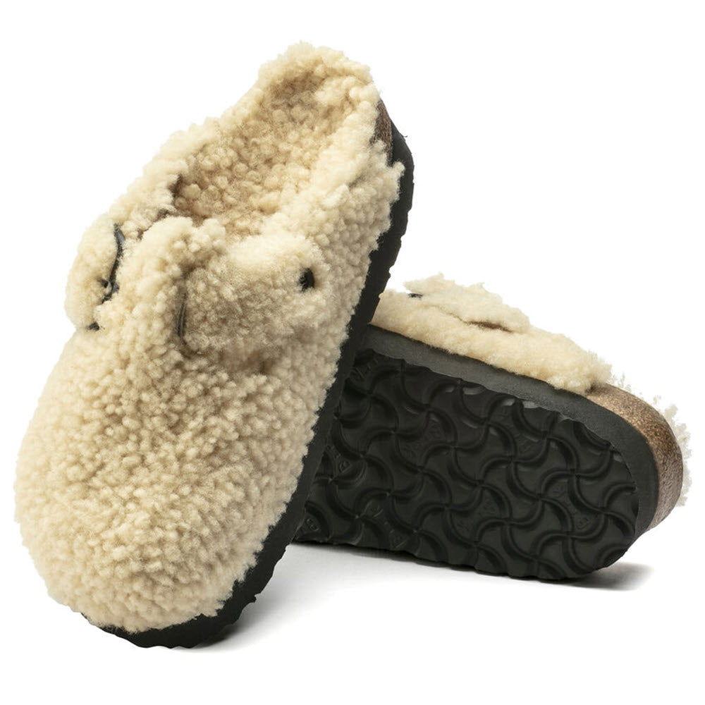 A pair of Birkenstock BOSTON PLATFORM BIG BUCKLE TEDDY EGGSHELL SHEARLING slip-on slippers with black rubber soles, featuring a beige, fleece-lined cozy Teddy style; one positioned upright and the other resting on its side.