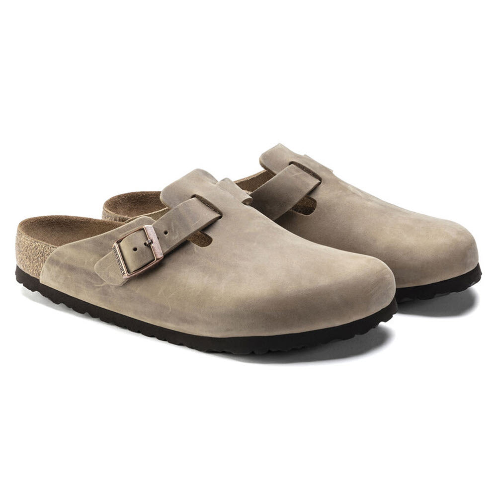 A pair of Birkenstock BIRKENSTOCK BOSTON TOBACCO LEATHER - ADULTS with adjustable buckles and cork footbeds, featuring soft black rubber soles.