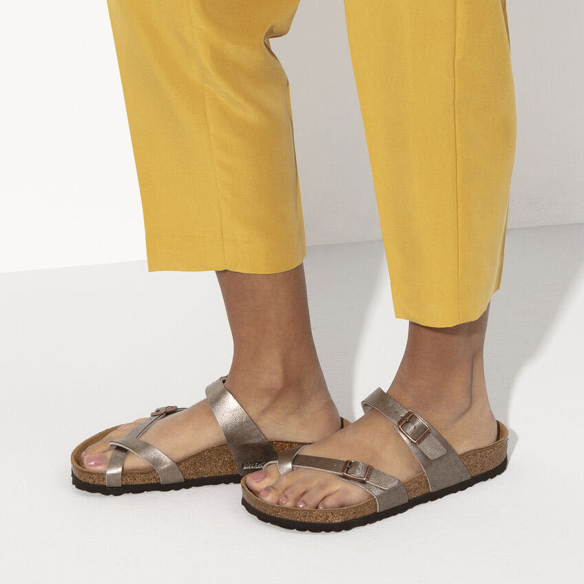 Person wearing yellow pants and bronze metallic Birkenstock BIRKENSTOCK MAYARI GRACEFUL TAUPE BIRKOFLOR - WOMENS sandals with adjustable straps, standing on a white surface.