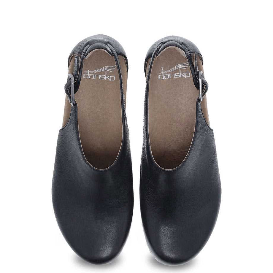 A pair of black closed-toe clogs with nubuck uppers and ankle straps viewed from above, branded with the Dansko logo on the insoles. This stylish transitional shoe, DANSKO SASSY BLACK - WOMENS by Dansko, features an EVA footbed for added comfort.