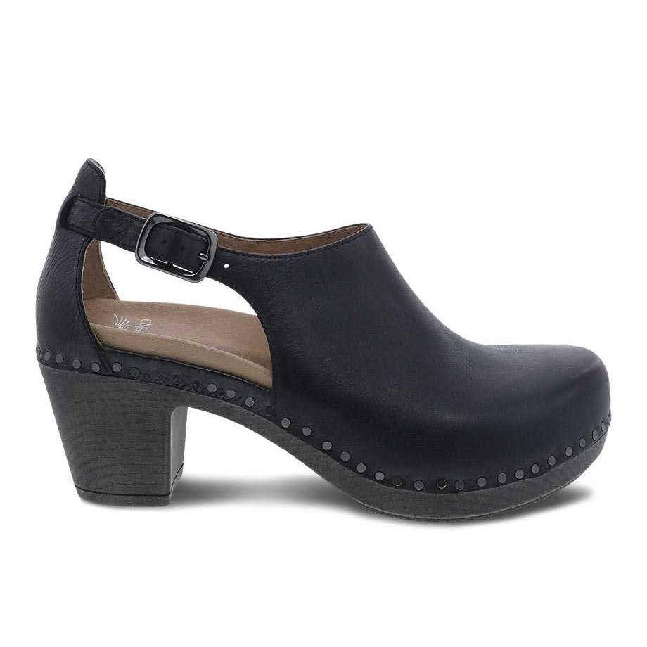 A side view of the Dansko DANSKO SASSY BLACK - WOMENS, a black leather, closed-toe clog with a chunky heel and a side buckle strap. Stud detailing is visible around the shoe&#39;s edge, making it an ideal transitional shoe.