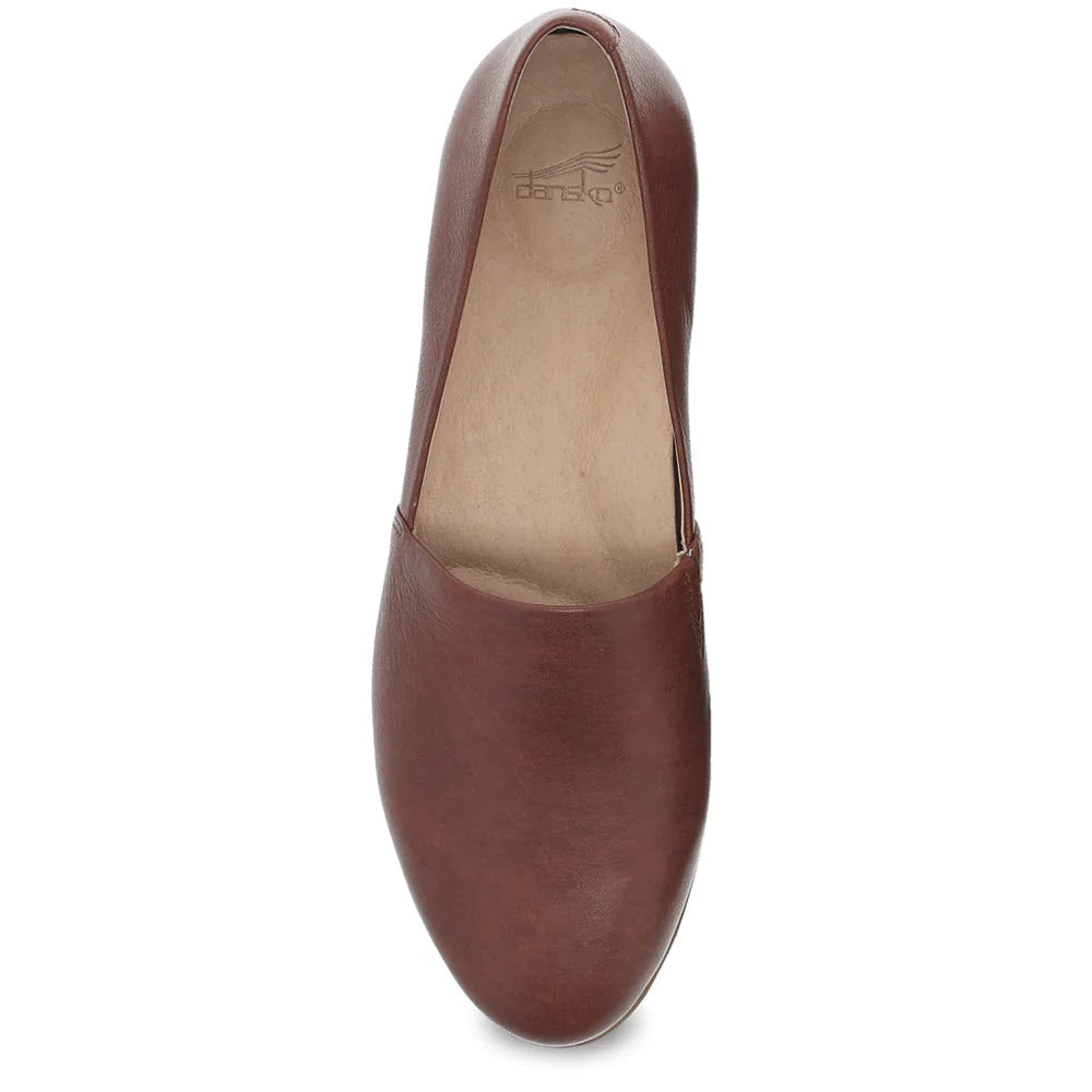 A top-down view of the Dansko Larisa Saddle for women showcases its single brown leather slip-on flat shoe, revealing a neutral beige insole with the Dansko logo visible inside. The design highlights the supportive arch and quality leather uppers.