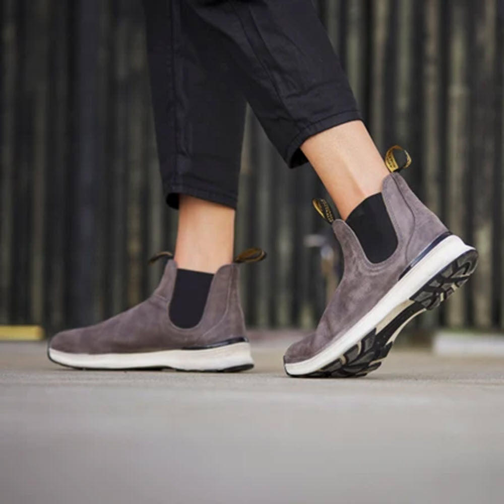 A person wearing BLUNDSTONE ACTIVE DUSTY GREY slip-on sneakers with black elastic panels from Blundstone walks on a concrete surface, enjoying the lightweight, breathable comfort and timeless look these shoes offer.