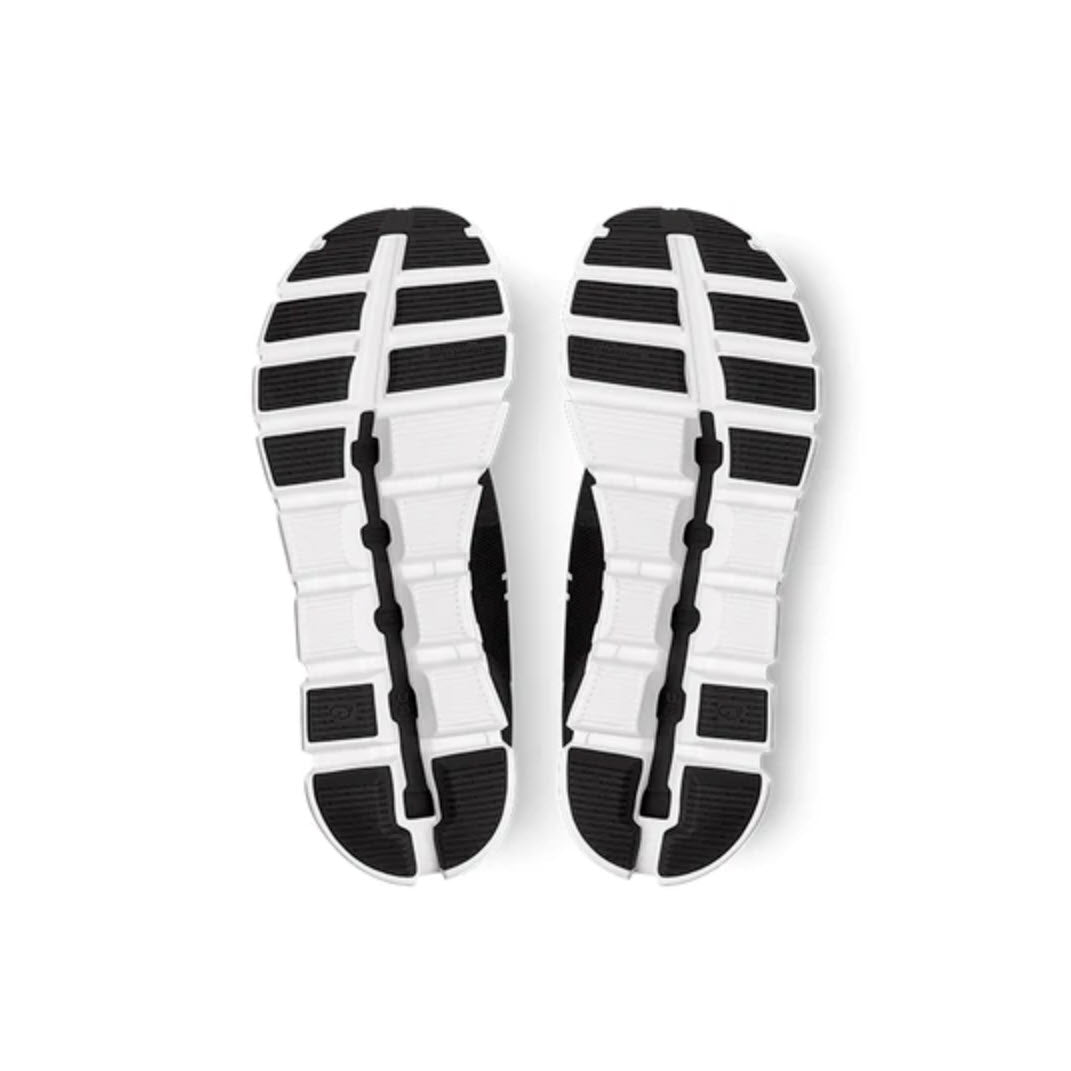 Bottom view of a pair of athletic shoes showing white soles with black grip patterns and segmented treads, featuring ON CLOUD 5 BLACK/WHITE - MENS enhanced by On Running&#39;s CloudTec® Zero-Gravity foam.