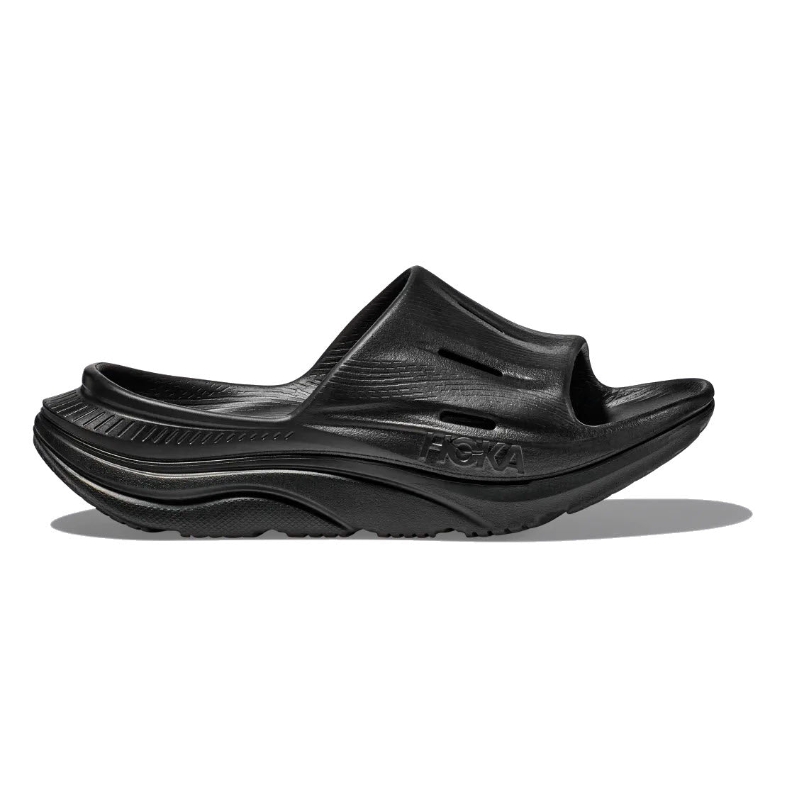 A HOKA ORA RECOVERY SLIDE 3 BLACK - ADULT, cushioned slip-on sandal with a thick, textured sole and open toe design. The brand name &quot;Hoka&quot; is embossed on the side, highlighting its comfort-driven style and foot-cradling comfort.
