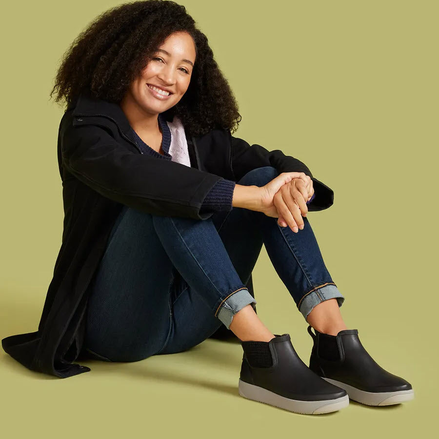 A woman sits on the floor against a green background, smiling, wearing a black coat, blue jeans, and functional Bogs Kicker Rain Chelsea II Black boots from Bogs.