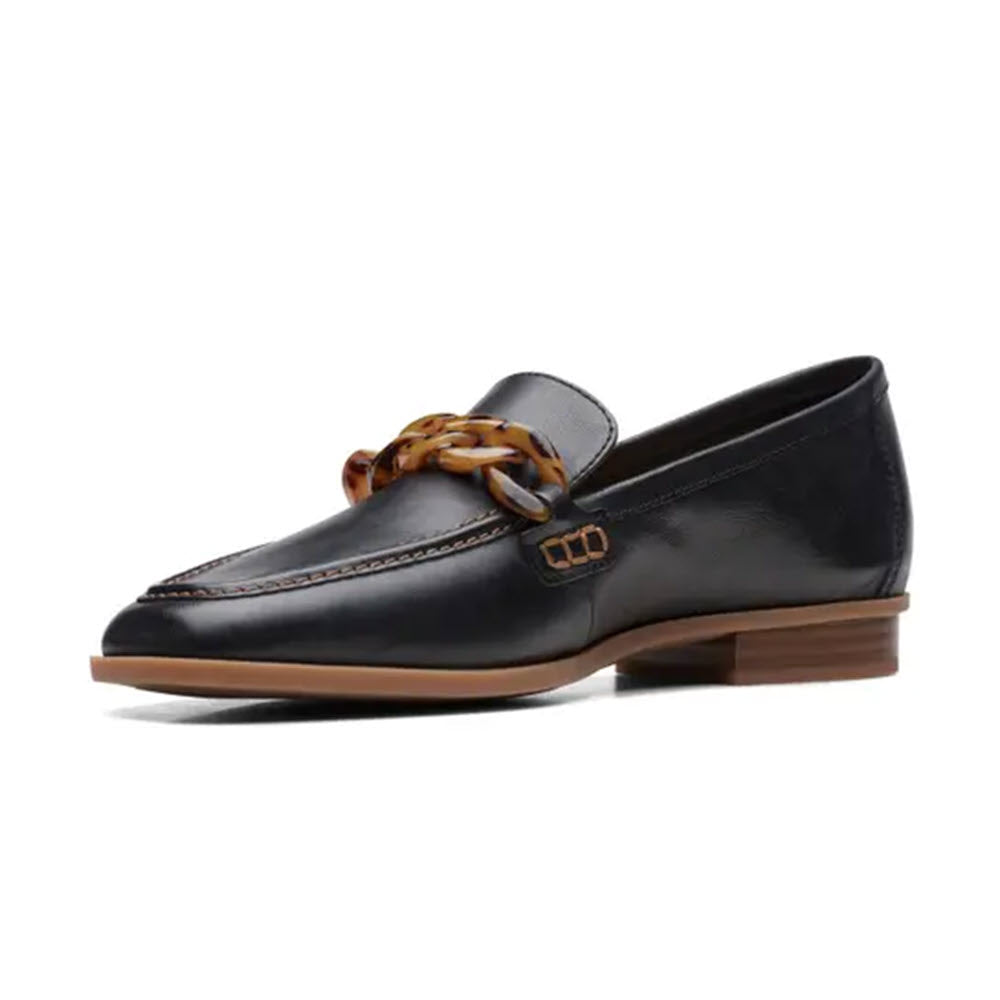 A single black leather CLARKS SARAFYNA IRIS BLACK - WOMENS loafer from Clarks, featuring a decorative vamp strap and a tan chain detail across the front, finished with a light brown sole, displayed on a white background.