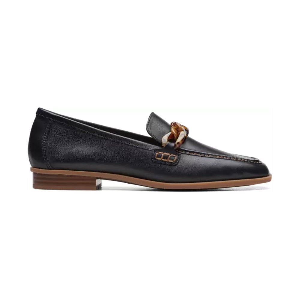 The CLARKS SARAFYNA IRIS BLACK - WOMENS loafer by Clarks showcases a black leather upper adorned with a decorative brown chain strap on the vamp, enhanced by a low wooden heel and brown stitching accents.