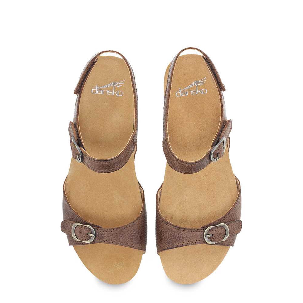 The Dansko Tricia Brown - Women&#39;s sandals are made from high-quality brown leather and feature ankle and toe straps, each adorned with a silver buckle. With its adjustable strap for premium comfort, this sandal is perfect for warm weather. The tan insole includes a logo printed near the heel.