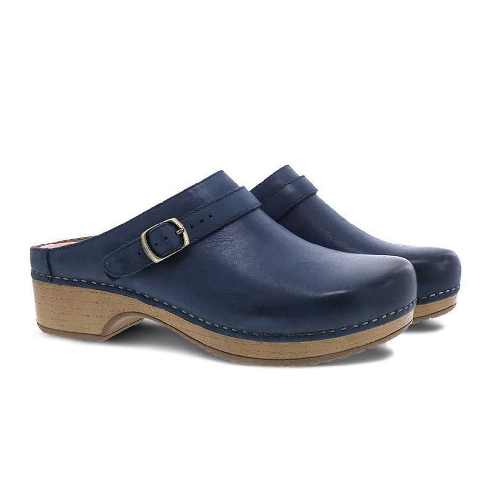 The Dansko Berry Navy Women&#39;s clogs feature navy blue nubuck uppers with stitching accents along the sides, wooden heels, and a buckle strap detail over the top.