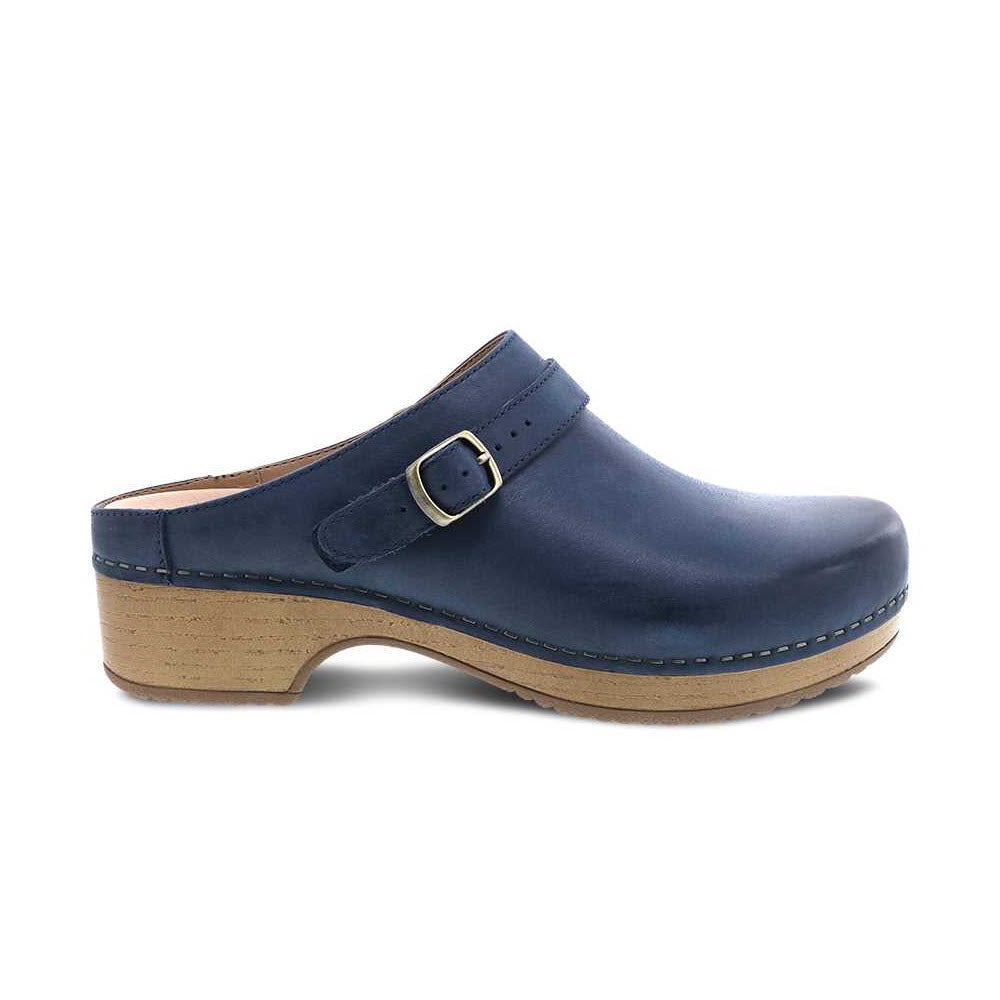 The Dansko Berry Navy - Women&#39;s is a navy blue leather mule from Dansko, featuring nubuck uppers and a wooden sole with a buckle strap across the top.