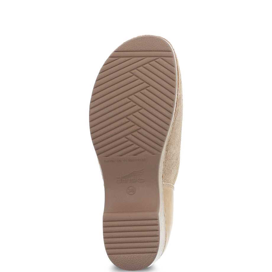 The image shows the bottom view of the DANSKO BRENNA SAND SUEDE - WOMENS, a single tan-colored casual slip-on shoe with a rubber sole. The sole features a herringbone tread pattern with the &quot;Dansko&quot; brand imprinted in the center, highlighting its stain resistance and odor control features.