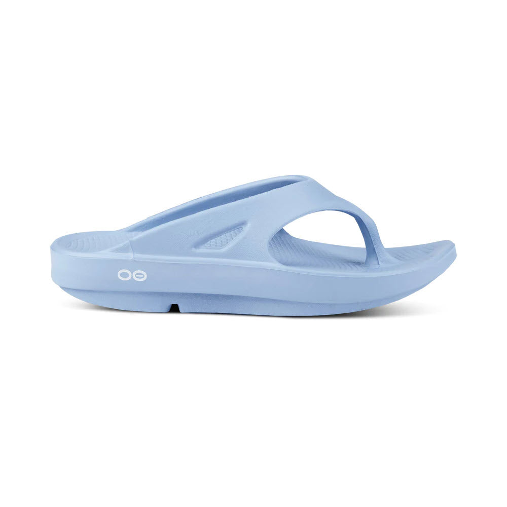 A light blue thong sandal with a single thick strap and a textured footbed, featuring OOfoam technology for ultimate comfort. The sandal has a slight platform heel and an &quot;OO&quot; logo on the side. The product is named OOFOS OORIGINAL THONG NEPTUNE BLUE - WOMENS by Oofos.