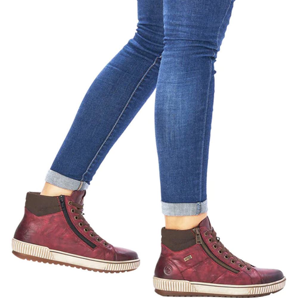 A person wearing rolled-up blue jeans and Remonte&#39;s City High Top Wine waterproof high-top sneakers, featuring dual side zippers, walking on a white background.