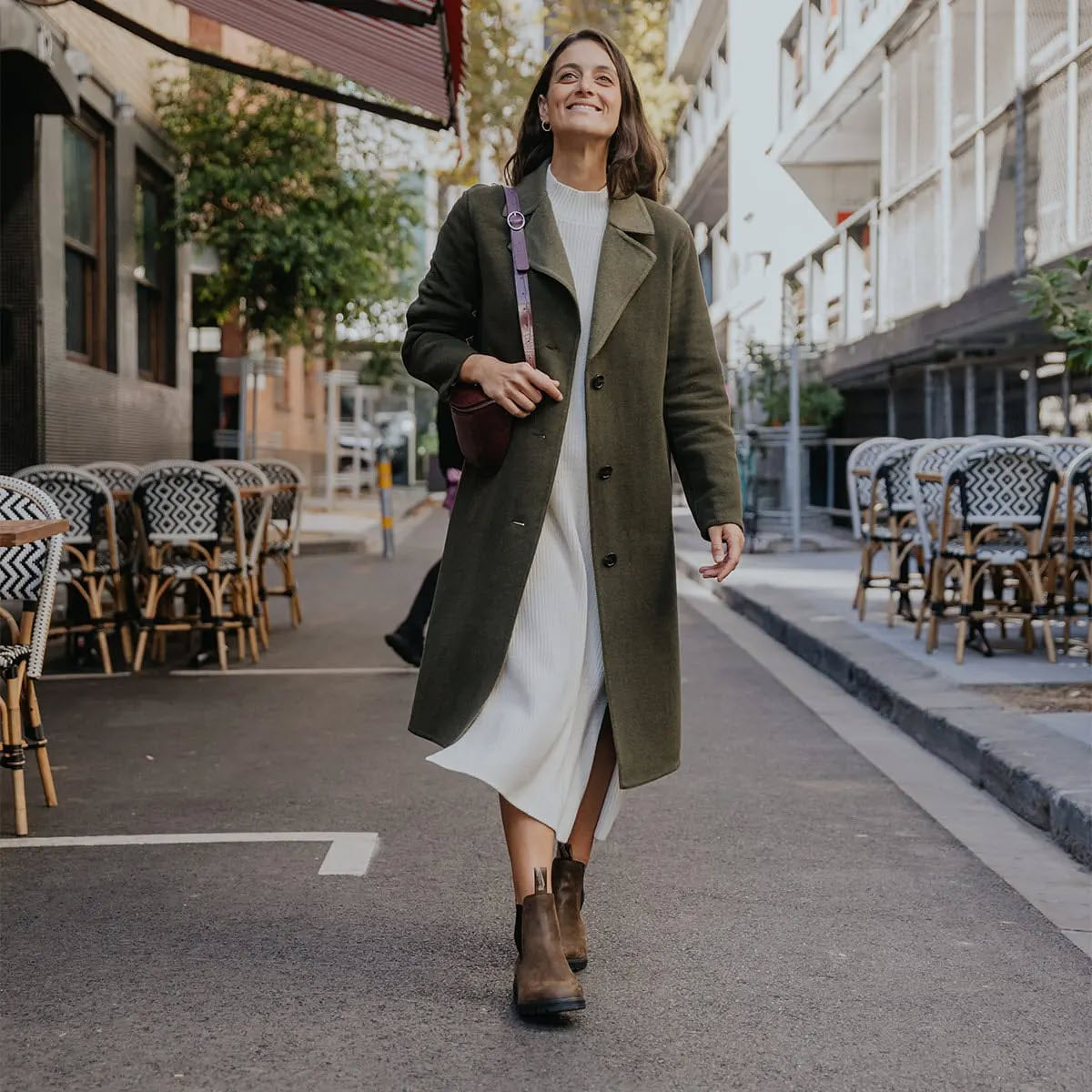 A woman in a long green coat and white dress walks down a street next to an outdoor dining area, her Blundstone BLUNDSTONE 2151 HIGH TOP ANTIQUE BROWN - WOMENS clicking softly on the cobblestones.