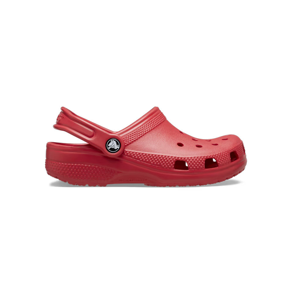 The CROCS CLASSIC CLOG VARSITY RED - KIDS from Crocs, a classic red rubber clog sandal with a perforated toe box, heel strap, and side ventilation holes, is shown on a plain white background. Featuring the signature Crocs Croslite material and a non-slip outsole, it offers both comfort and functionality.