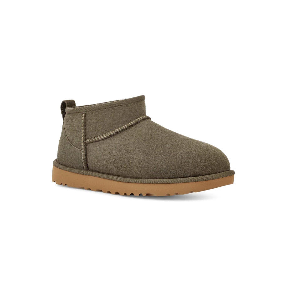 A single olive green ankle-high boot with a tan rubber sole and stitching details is shown against a white background, featuring the comfort of a sheepskin insole. The boot is the UGG CLASSIC ULTRA MINI FOREST NIGHT - WOMENS from Ugg.