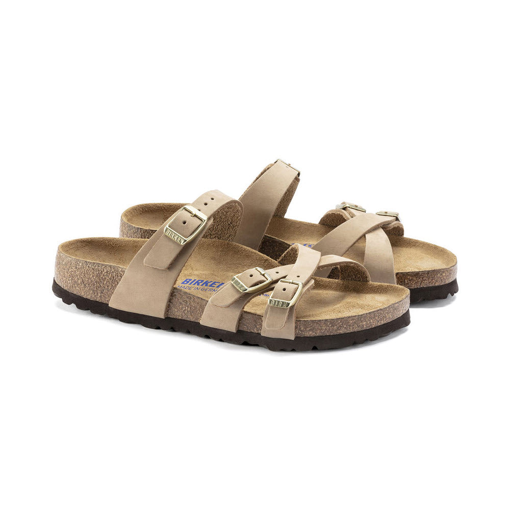 A pair of BIRKENSTOCK FRANCA SANDCASTLE NUBUCK - WOMENS with cork soles and stylish crisscrossed straps featuring metal buckles, complemented by a soft footbed for added comfort.