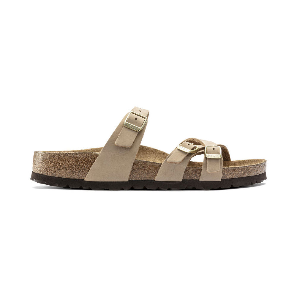 A beige Birkenstock BIRKENSTOCK FRANCA SANDCASTLE NUBUCK - WOMENS sandal with stylish crisscrossed nubuck leather straps and a cork sole. The sandal features a soft footbed and a rugged, textured black rubber outsole.