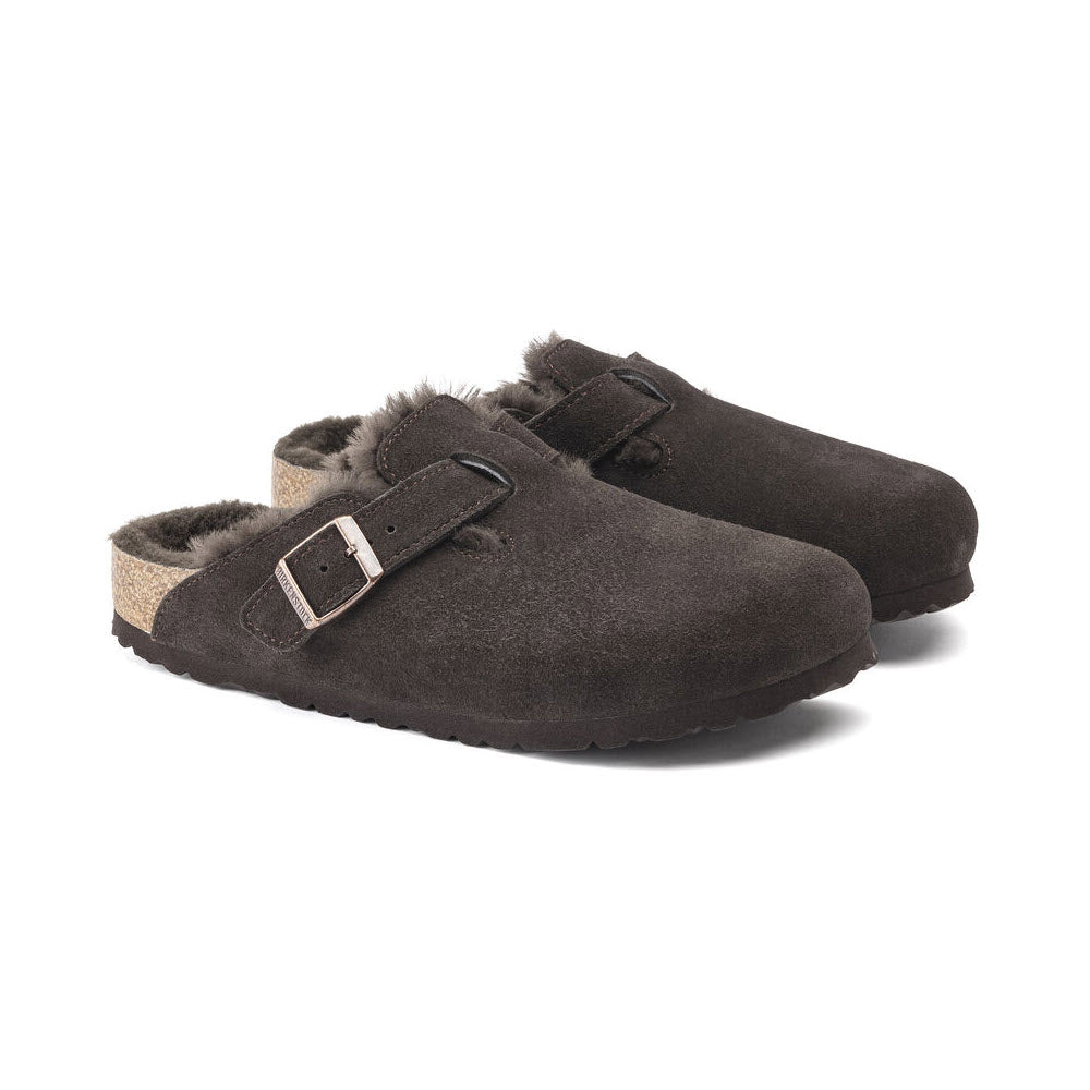 A pair of BIRKENSTOCK BOSTON SHEARLING MOCHA SUEDE - WOMENS from Birkenstock with shearling lining and an adjustable strap on each shoe, featuring a contoured cork footbed for added comfort, positioned on a white background. Perfect for casual-wear.