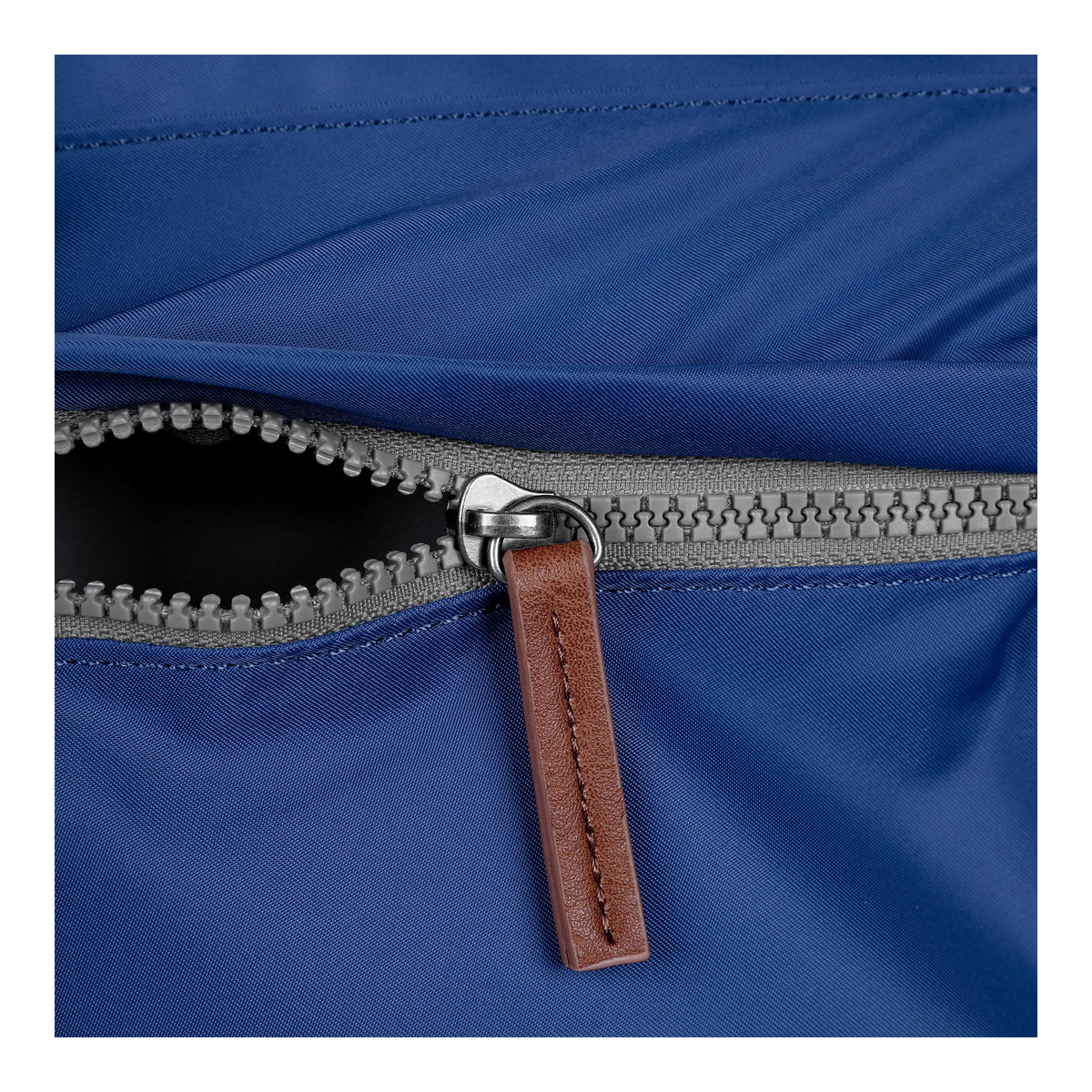 Close-up of a partially open zipper on a blue fabric, with a brown leather pull tab attached to the metal zipper slider, perfect for organizing your essentials in an ORI LONDON KENNINGTON SLING BAG BLUE zipped pocket from ORI LONDON.