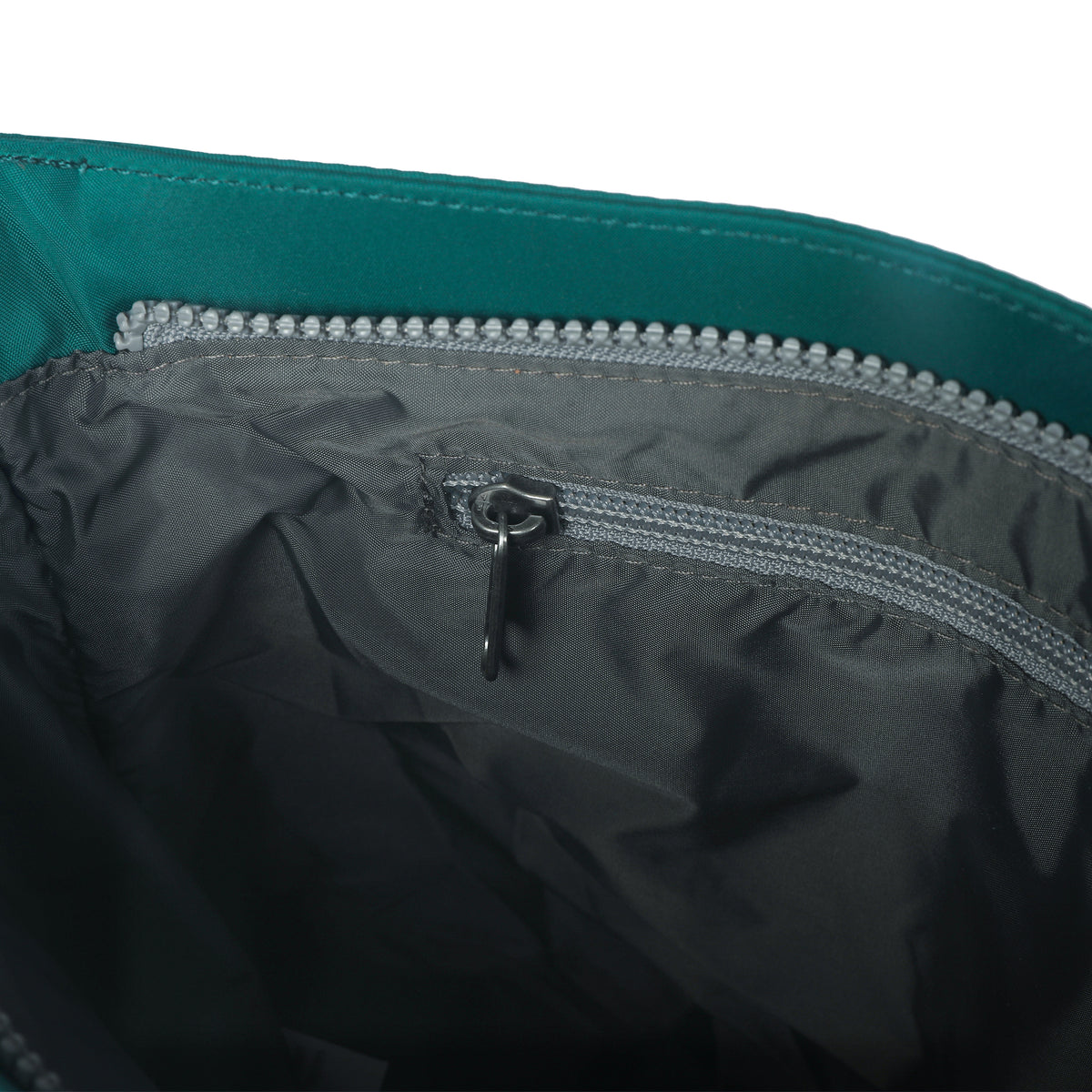 Close-up view of the interior of an Ori London ORI LONDON KENNINGTON CROSSBODY TEAL, showing a small gray zippered pocket inside a larger compartment. The bag is green with gray lining and made from recycled nylon.