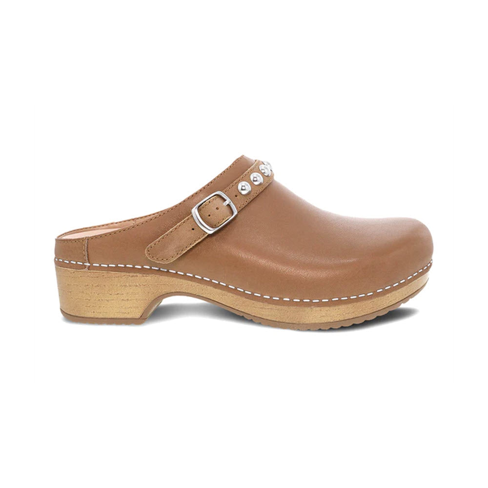 The Dansko Britton Tan women's mule, made from brown leather with a wooden sole, features a buckle strap and silver hardware decorative studs near the opening. It offers a versatile style perfect for any wardrobe.