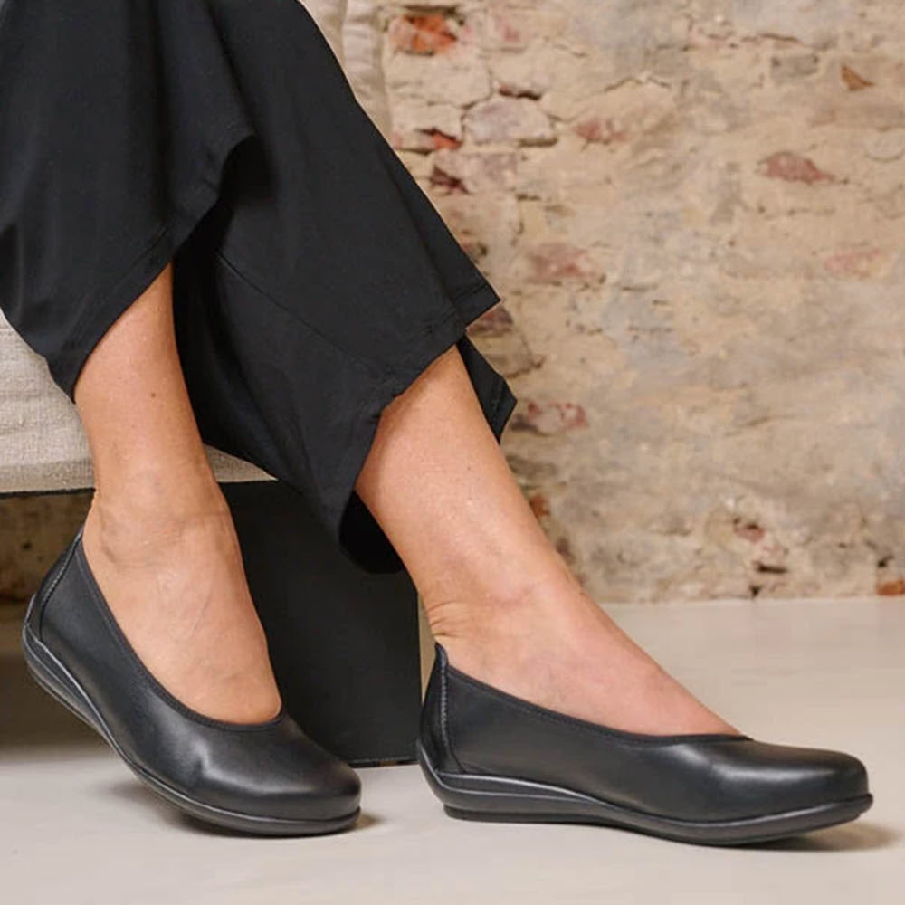 Person wearing black pants and black flat shoes, specifically the WOLKY DUNCAN F2F BLACK BIOCARE STRETCH - WOMENS from Wolky, seated against a backdrop of a rustic brick wall.