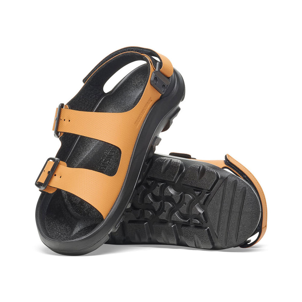 A pair of orange Birkenstock BIRKENSTOCK MOGAMI TERRA OASIS BURNT ORANGE - WOMENS with black, rugged PU soles and adjustable straps. One sandal is upright, while the other is on its side, showcasing the all-terrain grip tread pattern on the sole.