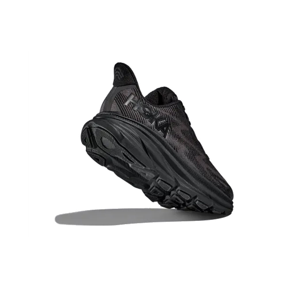 A HOKA CLIFTON 9 BLACK/BLACK - WOMENS athletic shoe features a thick sole and a breathable engineered mesh upper with visible tread pattern. Positioned at an angle, it casts a shadow underneath.