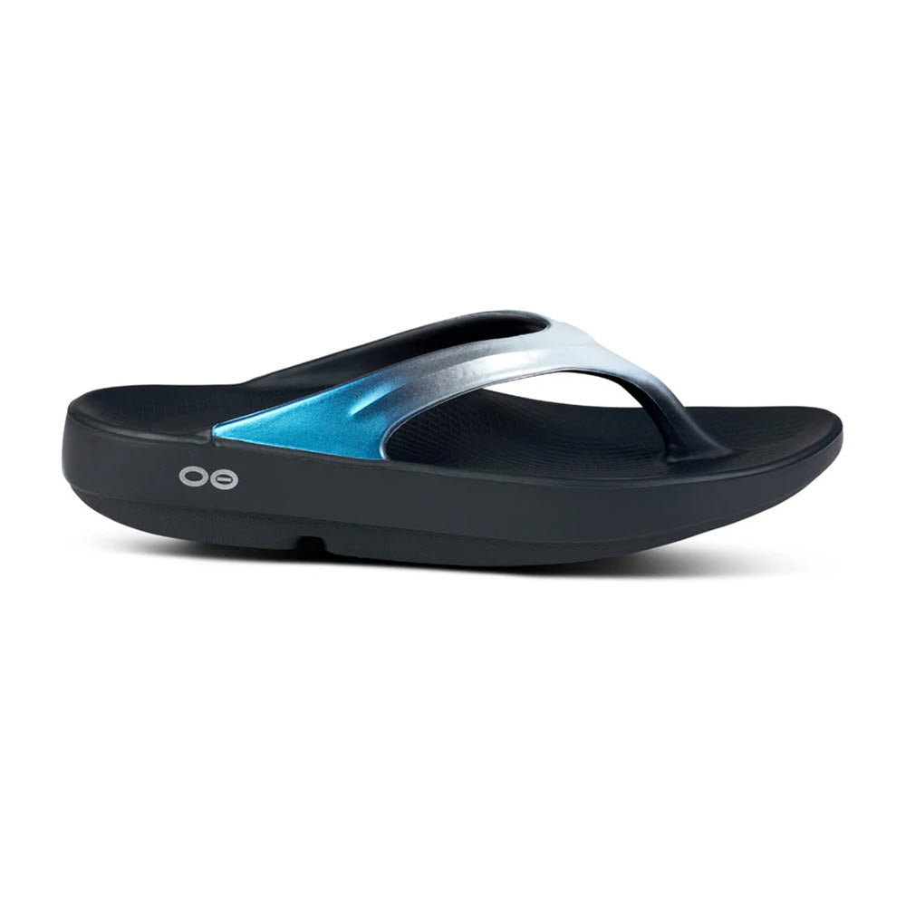 The OOFOS OOLALA LUXE CALYPSO - WOMENS by Oofos is a single black flip-flop sandal featuring a blue and gray gradient strap. It boasts cushioned soles, a curved design, and OOfoam footbed cushioning for added comfort. Made from moisture and odor-resistant materials, this sandal is perfect for any activity.