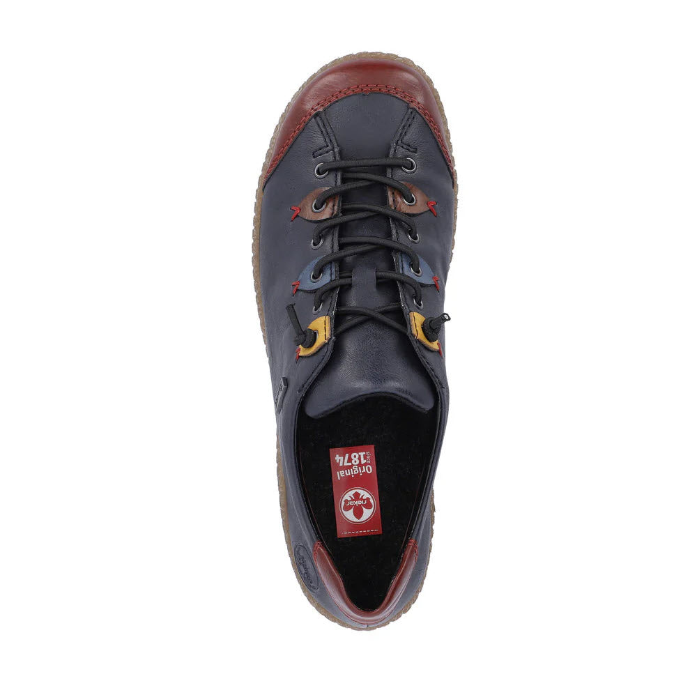 A top-down view of the RIEKER EURO CITY SLIP ON OXFORD NAVY MULTI - WOMENS, showcasing its navy blue and brown design with black laces and a visible red and white Rieker brand logo inside. This casual shoe also features a removable insole for added comfort.