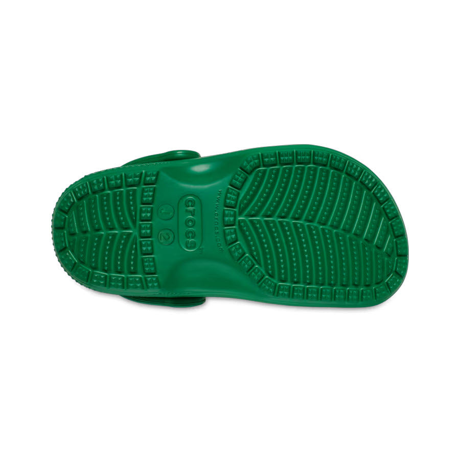 The image showcases the bottom sole of a green kids&#39; CROCS CLASSIC CLOG GREEN IVY from Crocs, featuring a textured, slip-resistant tread pattern for added comfort and support.